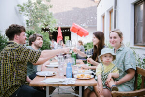 Eine Gruppe junger Menschen genießt Wein und Essen auf einem großen Heurigentisch und hat Spaß dabei. 