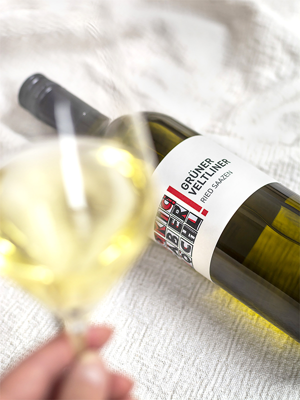 Eine Flasche Grüner Veltliner Ried Saazen vom Weingut Faber-Köchl liegend auf einer weißen Decke, davor hält jemand ein Glas Weißwein in der Hand