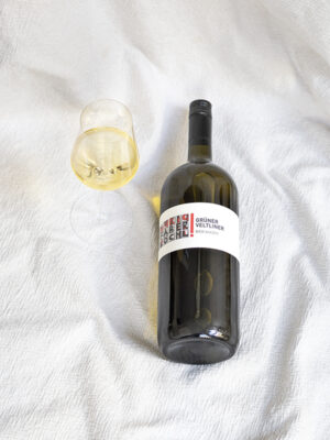 Magnum-Flasche des Grüner Veltliner Saazen liegt auf weißem Tuch, daneben ein Achterl-Glas mit Weißwein